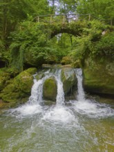 An idyllic waterfall flows under a rustic bridge, surrounded by lush greenery and rocks, aerial