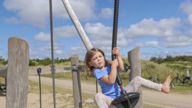 A child happily jumps on a zip wire in a wide, sunny playground setting with a cloudy, blue sky