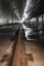 Long, damp tunnel made of concrete with rails and side lighting, looks gloomy and industrial,