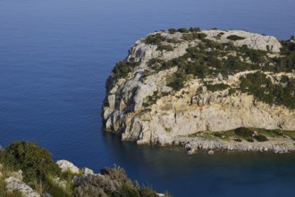 Steep rocky coast with calm, blue water and green vegetation under a clear sky, Anthony Quinn Bay,