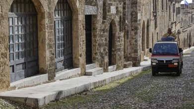 An old building with large windows along a cobblestone street and a moving small flatbed truck,