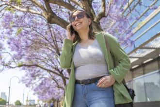 A young woman in casual attire talks on her smartphone while sightseeing by jacaranda trees and a