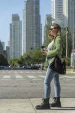 Young woman is holding a reusable coffee cup and waiting to cross the street in a big city with