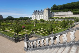 A castle with a magnificent, symmetrical garden full of flowers and shrubs under a clear blue sky,