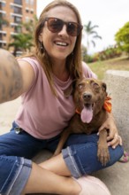 A smiling woman wearing sunglasses sits on the ground, taking a selfie while petting her dog, who