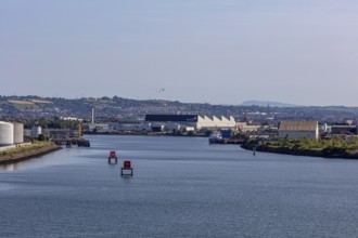 River landscape with industrial buildings, buoys and a flying aeroplane under a blue sky, Belfast