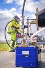 Worker in protective equipment operates outdoor machine with hoses and metal parts, glass fibre