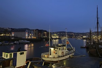 Fishing boats in the harbour at night with glowing city lights in the background, Nidarelva,