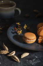 Orange macarons or macaroons cakes with cup of coffee on a black concrete background. Hard light,