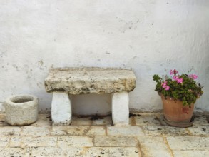 Stone bench and flower pot with geraniums on a rustic stone floor in front of a white wall, Apulia,