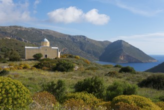 Small church by the sea with mountain backdrop under blue sky, surrounded by green landscape,