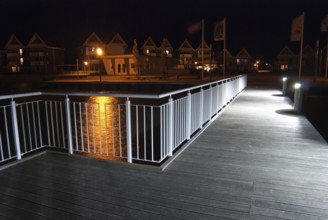 Pier with illuminated lamps, railings and light reflections in the water at night, flags and houses