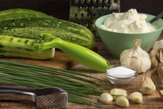 Fresh ingredients such as cucumbers, garlic, chives and sour cream on a wooden table with a peeler