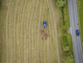 Aerial view of a tractor with hay tedder on a hay field next to a country road, surrounded by