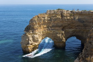 A boat sails under a natural rock arch along an impressive rocky coast, Praia da Marinha, double