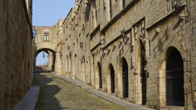 A historic alleyway with stone buildings and arches, blue and white flags in the background,