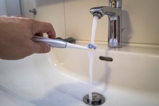 A person holds an electric toothbrush under a running tap in the bathroom, symbolic image of water