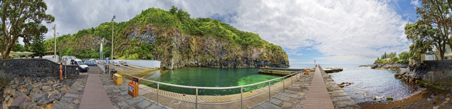 360° view, wooden walkway leading to green rocky coast and clear ocean under blue sky with clouds,