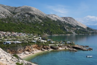 Coastal landscape with rocky mountains and clear blue water, lined with small houses under a sunny