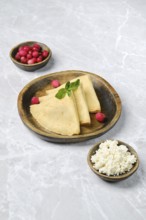 Three golden crepes are elegantly arranged on a wooden plate, accompanied by small bowls of fresh