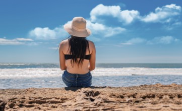 Girl in hat sitting with her back facing the sea, Young woman in hat sitting with her back looking