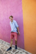 Young man leaning against a pink and orange house wall, colourful houses on the island of Burano,