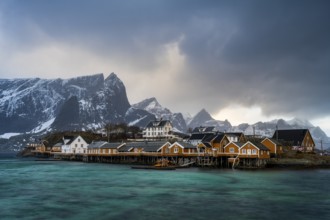 Traditional yellow rorbuer cabins on Sakrisøy Island, snow-capped rocky mountains in the back,