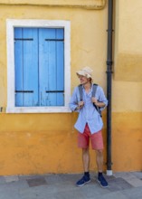 Young man with backpack in front of yellow house wall, yellow house facade with windows with blue