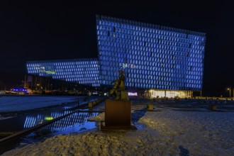 Bronze statue of the man with the cello, behind it the illuminated facade of the concert hall and