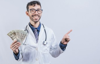 Happy young doctor holding money and pointing to side. Handsome smiling doctor showing money and