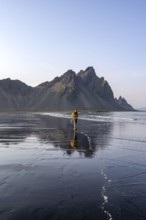 Tourist at black lava beach, sandy beach and sea, mountains Klifatindur reflected in the water,