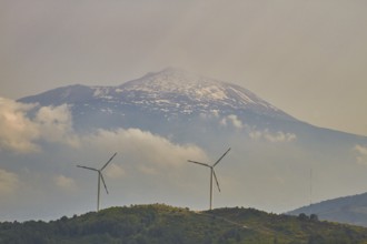 Two wind generators on a green hill, snow-capped Mount Etna in the background, Nebrodi National