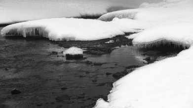 Snow mounds and icicles in the Skoga River below the Skogafoss waterfall, black and white photo,