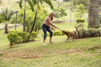 Middle-aged woman wearing sunglasses, playing with her playful dog in a grassy area, as the dog