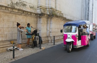 A violinist and a singer play music in front of an old wall while a pink tuk-tuk drives by, Old