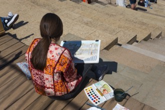 Woman painting with watercolours on the steps of a Roman theatre, surrounded by painting tools and