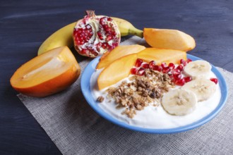 A blue plate with greek yogurt, granola, persimmon, banana, pomegranate black wooden background.