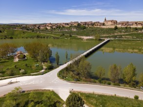 A village along a river with a bridge and a road, surrounded by green landscape and trees under a