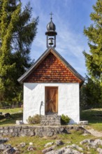 Small chapel made of wood and stone surrounded by tall trees under a clear sky, Winklmoosalm