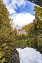 A river flows through an autumnal forest landscape with a hotel in the background under a cloudy