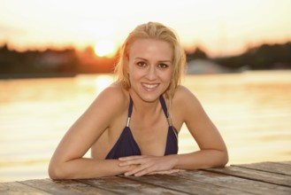 Woman in a bikini leaning on a dock with a sunset background, smiling relaxedly, Austria, Europe