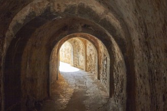 View through a dark tunnel with arches and stone walls leading to a light-coloured exterior