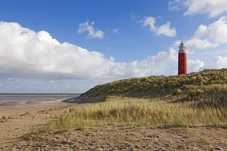 A red lighthouse stands on a grassy dune on the beach under a blue sky with clouds, Eierland