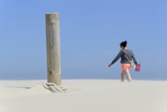 Boundary post on the beach, rope blown over by sand, person crossing the sand drifts, blue sky,