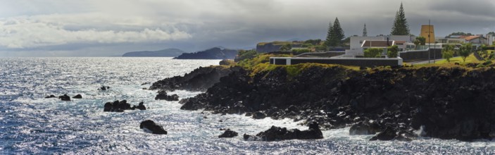 Coast with strong surf, rocky shore and some houses on a cliff under cloudy sky, Capelas, Sao