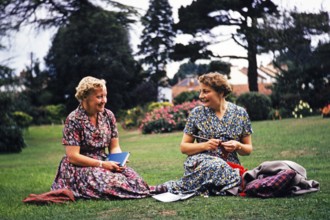 Two women friends sitting on graß lawn one knitting, smiling at each other, Dawlish, Devon,