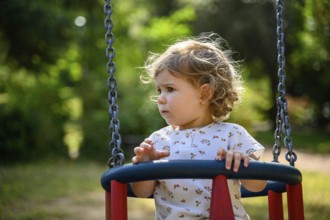 Child, 2 years old, on a swing outdoors, surrounded by green nature in sunny weather