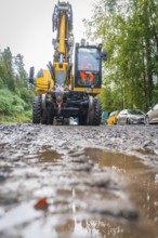 A yellow excavator works on a muddy road in the forest with two construction workers in sight,