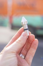 Hand holding a small metal component in front of a blurred background, Wend Glasfasermontage during