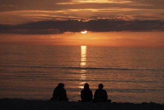 Three people sitting on the beach watching the sunset over the sea, silhouettes in front of warm